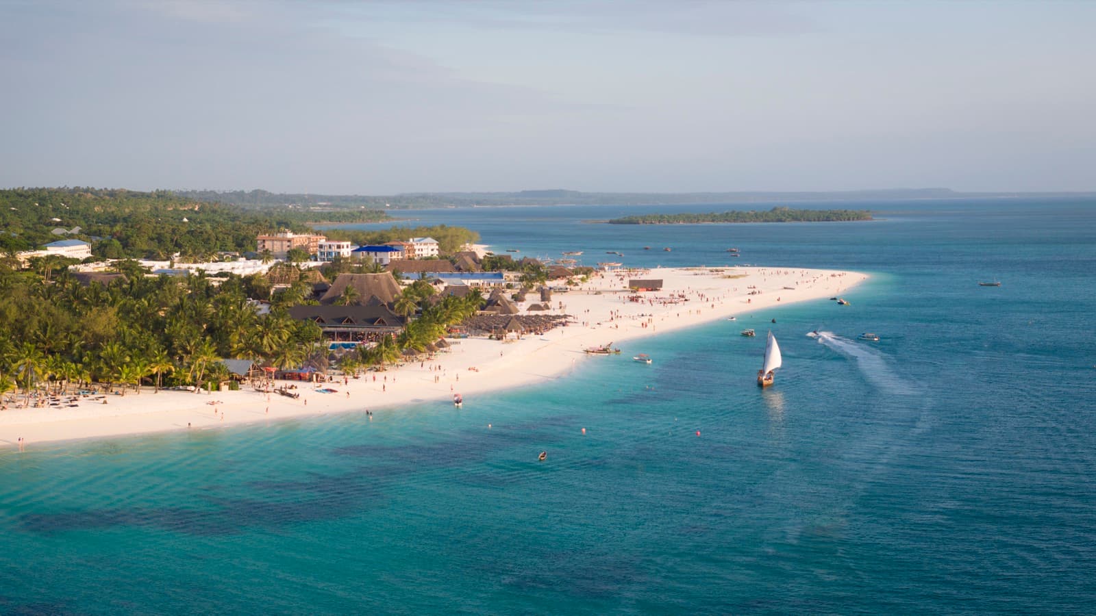 A dhow under sail off a long Zanzibar beach, with palms and low resort buildings along the shore